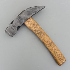 May include: A vintage metal hammer with a wooden handle. The hammer head is dark grey and has the letters "S.M.&" stamped on the side. The handle is made of light brown wood with a visible grain pattern.