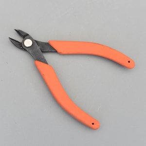 以下が含まれることがあります： オレンジ色のハンドルと黒い刃が付いたワイヤーカッターのクローズアップ。 このツールは、ワイヤーや小さなオブジェクトを切断するように設計されています。