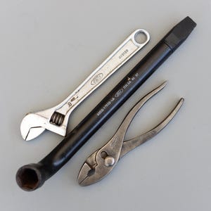 Puede incluir: Un surtido de herramientas, que incluye una llave inglesa ajustable plateada, una llave de tuercas negra y unos alicates de junta deslizante plateados. La llave inglesa ajustable tiene el logotipo de Ford y el número 8. La llave de tuercas tiene el logotipo de Ford y el texto "93EB-17032-A".