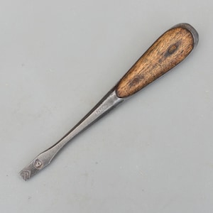 Puede incluir: Un cincel antiguo con mango de madera y hoja de metal. El mango es de color marrón claro y la hoja es de un gris más oscuro. El cincel parece estar bien usado, con algo de desgaste visible.