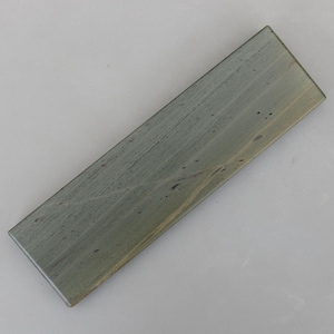 Puede incluir: Una piedra de afilar rectangular con un tono gris verdoso. La piedra tiene una superficie ligeramente texturizada y patrones de grano visibles. Está diseñada para afilar cuchillos y otras herramientas de corte.