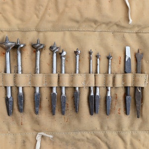 May include: A set of 12 drill bits in a canvas roll-up case. The drill bits are made of metal and have different sizes. The case is brown and has a white strap.