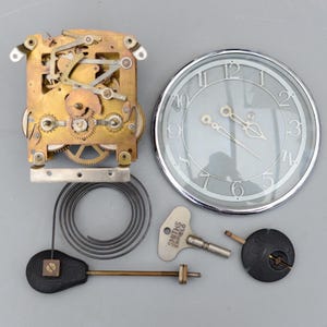 May include: Clock parts arranged on a gray surface. Components include a clock face with silver hands, a brass movement, a coiled spring, a pendulum, and a key with "SMITHS ENFIELD" text.