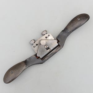 May include: A vintage hand-held spokeshave tool with a dark gray finish. The tool features a curved blade, two adjustment screws, and textured handles for grip. The spokeshave is designed for shaping wood.