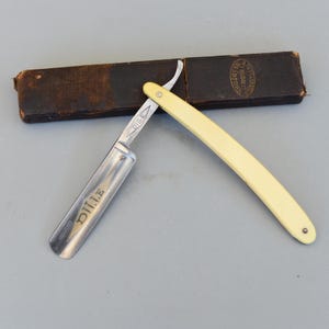 Puede incluir: Una navaja de afeitar abierta con mango color crema y hoja plateada. La hoja está marcada con la palabra "DILE". En el fondo, un estuche de cuero marrón oscuro. Una herramienta de afeitado vintage.