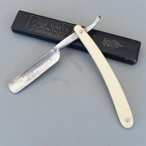 以下が含まれることがあります： クリーム色のハンドルと銀色の刃が付いたオープンストレートカミソリ。黒いケースの隣にあります。刃には「Sheffield England John Watts」と刻まれています。ケースには「John Watts Sheffield England Established 1765」とも書かれています。