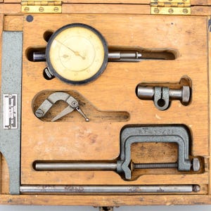 Op de afbeelding: Een houten kist met een wijzerplaatmeter, een klem, een staaf en een klein gereedschap. De wijzerplaatmeter heeft een zwarte wijzerplaat met witte markeringen en een zilveren naald. De klem is van metaal en heeft een zwarte handgreep. De staaf is van metaal en is lang en dun. Het kleine gereedschap is van metaal en heeft een gebogen handgreep.