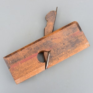 May include: A wooden hand plane with a metal blade and a red painted stripe. The plane is made of wood and has a worn, rustic look.