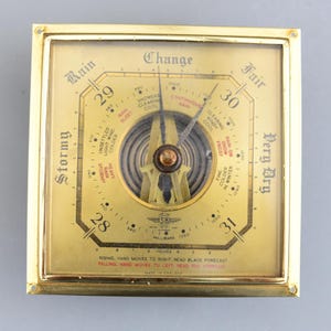 May include: A vintage square barometer with a gold-coloured frame and a brass face. The face features a needle, numbers, and text indicating weather conditions like "Stormy" and "Fair." The barometer measures in inches and millibars.