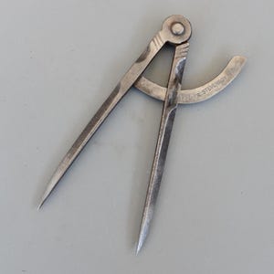 May include: A vintage metal compass with pointed tips and a curved arm. The compass is silver-toned and shows signs of wear. The text "R. STEVENSON" is visible on the curved arm. This tool is likely used for measuring or drawing circles.