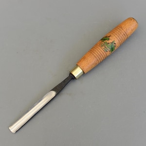 Puede incluir: Un formón de madera con un mango de madera marrón claro y una hoja de metal. El mango tiene un logotipo verde y blanco que dice "Taylor Tools". El formón está diseñado para carpintería y tallado.