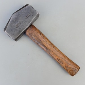 May include: A vintage metal hammer with a rectangular head and a wooden handle. The hammer head is dark gray with the text "KEARNS WASHINGTON CO" and "4 LBS" stamped on it. The handle is made of light brown wood.