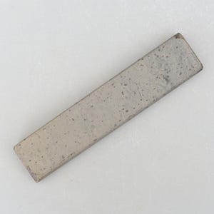 Könnte beinhalten: Eine lange, rechteckige, graue und weiße Keramikfliese mit rauer Textur. Die Fliese hat ein dezentes Muster aus kleinen, dunkelgrauen Flecken.
