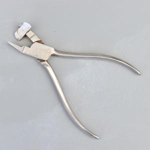 May include: Silver metal pliers with a curved handle and a flat jaw with a small, square-shaped head.