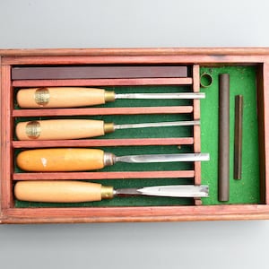 Könnte beinhalten: Eine Holzkiste mit grünem Filzbezug enthält einen Satz von fünf Holzschnitzwerkzeugen mit Holzgriffen. Die Werkzeuge sind in einer Reihe angeordnet, wobei die Griffe nach oben zeigen. Die Kiste enthält außerdem einen Schleifstein und eine Metallstange.