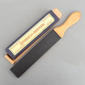 以下が含まれることがあります： 木製のハンドルと黒い革のストロップが付いたレザーストロップ。箱は青で、金色の文字で「RAZOR-STROP STREICH-RIEMEN - CUIR A RASOIR - ASENTATOR - CORAME」と書かれています。