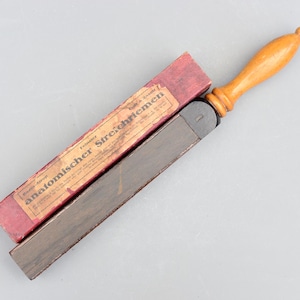 以下が含まれることがあります： 赤い箱に入ったヴィンテージの木製レザーストロープ。箱にはドイツ語で「feiner anatomischer Strichriemen」と書いてあり、これは「細かい解剖学的ストロープ」という意味です。