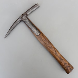 May include: A vintage pick hammer. It features a dark wooden handle and a metal head. The head has a pointed pick and a flat striking surface. The metal shows signs of wear and tear.