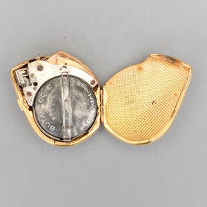 May include: Open gold-coloured compact with a battery and mechanical components visible. The interior of the compact is gold, with a textured pattern on the lid. The battery is dark grey with white text. The compact is open, revealing its inner workings.