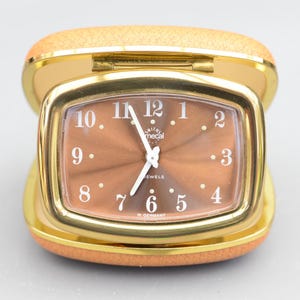 Puede incluir: Un reloj despertador de viaje dorado con una esfera marrón y números blancos. El reloj tiene el logotipo de Smiths Mecal y dice "7 Jewels" y "W. Germany".