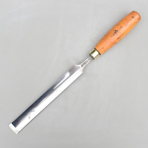 May include: A woodworking chisel with a polished steel blade and a wooden handle. The handle is a light brown color with a brass ferrule. The blade is rectangular and tapers to a sharp edge. The chisel is designed for shaping wood.