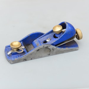May include: A small, blue hand plane with brass adjustment knobs. The plane has a metal blade and a blue painted body, showing signs of wear. The tool is designed for woodworking and smoothing surfaces.