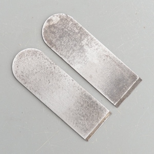 May include: Two silver-coloured metal blades for a wood plane. The blades are slightly curved and have a bevelled edge.