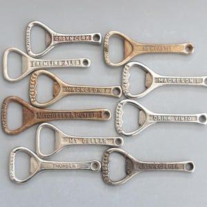 Puede incluir: Colección de abridores de botellas vintage en tonos plateados y bronce. Cada abridor presenta el nombre de una marca diferente, como "Crown Cork", "Mackeson" y "Drink Vimto". Los abridores están dispuestos sobre una superficie gris.