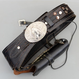 May include: Black leather gun belt with a silver buckle depicting a horse head. The belt has decorative stitching and metal studs. A black leather holster is attached. This is a western-style accessory.