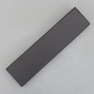 May include: A rectangular, matte black object, possibly a box or a component, is positioned diagonally against a light gray background. The object's surface appears smooth and uniform, with a subtle sheen. The overall aesthetic is minimalist and modern.