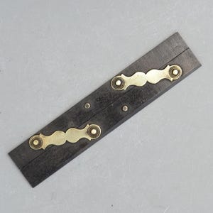 May include: A black wooden folding rule with brass accents. The rule has two brass hinges and two brass plates with decorative edges. The rule is closed and lying flat on a grey surface.