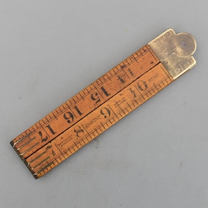 Peut inclure: Une règle pliante en bois vintage avec des raccords en laiton. La règle est marquée de chiffres noirs et de lignes de mesure. Le texte "RABONE" et "MADE IN ENGLAND" sont visibles. La règle mesure environ 43 cm de long lorsqu'elle est entièrement déployée.