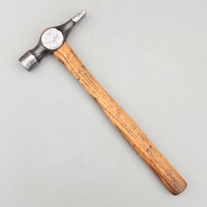 May include: A metal hammer with a wooden handle. The hammerhead is silver and has a flat face on one side and a pointed tip on the other. The handle is made of wood and is a light brown color.