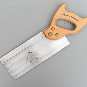 Peut inclure: Une scie à tenon Spear & Jackson vintage avec une poignée en bois et des accents en laiton. La lame de scie est argentée et présente une légère patine.