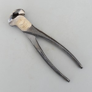 以下が含まれることがあります： ヴィンテージのクレセントニッパー。銀色のヘッドと濃い灰色のハンドルが付いた手工具です。この工具は切断と把持用に設計されており、ブランド名と「U.S.A.」がヘッドに刻印されています。