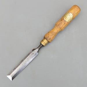 May include: A woodworking chisel with a wooden handle and a metal blade. The handle is light brown with a brass ferrule. The blade is silver and angled at the end. A gold-colored label is on the handle.