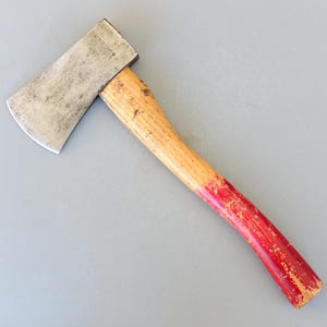以下が含まれることがあります： 赤い塗装が施された木製の柄を持つビンテージの斧。斧の頭は金属製で、わずかに緑青が付着している。