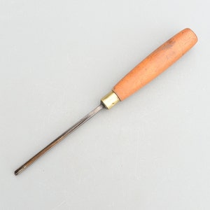 Può includere: Uno strumento per intagliare il legno con un manico in legno e una lama in metallo. Il manico è di colore marrone chiaro e la lama è di colore grigio scuro.