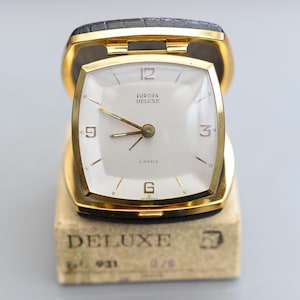 May include: A vintage Europa Deluxe travel clock in a gold-tone case with a white face. The clock features black hands and hour markers, with the text "EUROPA DELUXE" and "2 JEWELS". It's housed in a black leather case and rests on a cardboard box.