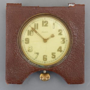 Pode incluir: Um relógio vintage com caixa de couro castanho e mostrador creme. O mostrador apresenta algarismos e ponteiros pretos, com o texto "8 DAYS" acima do centro. Um botão dourado pende por baixo do relógio. O relógio é quadrado com bordas arredondadas.