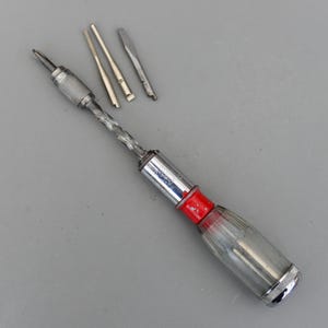 May include: A vintage screwdriver set with a clear plastic handle, red accent, and silver metal components. The set includes the main screwdriver and several interchangeable bits. The handle is approximately 15 cm long.