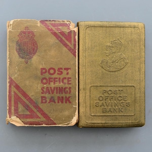 May include: A vintage Post Office Savings Bank book and case. The book has a worn, tan cover with red accents and text. The gold-coloured case features an embossed image and text. Both items are rectangular.