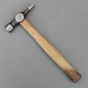 Peut inclure: Un marteau en métal avec un manche en bois. Le marteau a une tête arrondie d'un côté et une pointe de l'autre. Le manche est brun clair et la tête du marteau est argentée.
