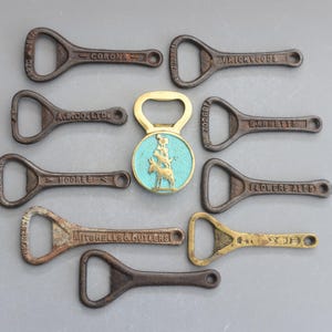 Może przedstawiać: Kolekcja dziesięciu zabytkowych otwieraczy do butelek, w większości wykonanych z metalu z patyną rdzy. Otwieracze mają różne nazwy marek, w tym Corona, A.W. Rooke Ltd, Moores, Garretts, Flowers Ales i Mitchells & Butlers. Jeden otwieracz jest wykonany z mosiądzu i ma wzór osła z niebieskiego emaliowanego.