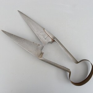 May include: Antique metal shears with sharp, pointed blades and a loop handle. The shears have a weathered appearance, with markings visible on the blades. The blades are open, revealing their cutting edges. The shears are probably for cutting fabric or similar materials.