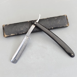 May include: A vintage straight razor with a black handle and a silver blade, alongside a black leather case. The blade is marked with the text "FRISEUR FREUND NO. 1". A classic grooming tool.