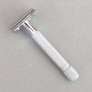 May include: A chrome safety razor with a textured handle. The razor head is rectangular, and the handle is cylindrical with a diamond pattern for grip. The razor is designed for shaving.
