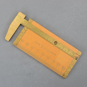 May include: A vintage wooden and brass caliper tool. The tool features a wooden body with an orange hue and brass measuring scales. The caliper is marked with numbers and measurements in centimetres.
