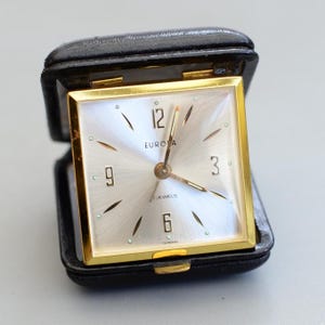 Peut inclure: Un réveil de voyage Europa vintage dans un étui en cuir noir. Le cadran de l'horloge est argenté avec des aiguilles dorées et un cadre doré. L'horloge affiche les chiffres 3, 6, 9 et 12, avec le mot "EUROPA" au-dessus du centre.