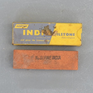 Könnte beinhalten: Eine gelbe und blaue Schachtel mit dem Text "Norton India Oilstone Made in England" und dem Text "still gives the keenest edge". Neben der Schachtel liegt ein rotbrauner Schleifstein mit dem Text "No. 61 Fine India".